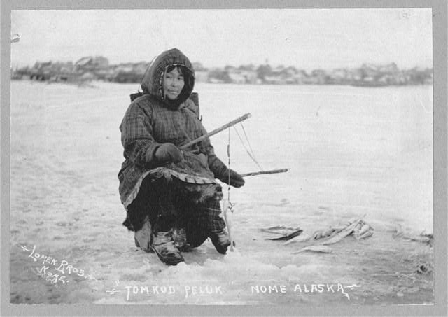 Black and white photograph of an Eskimo woman holding two rods for ice fishing and smiling slightly at the camera. She has caught a fish. 